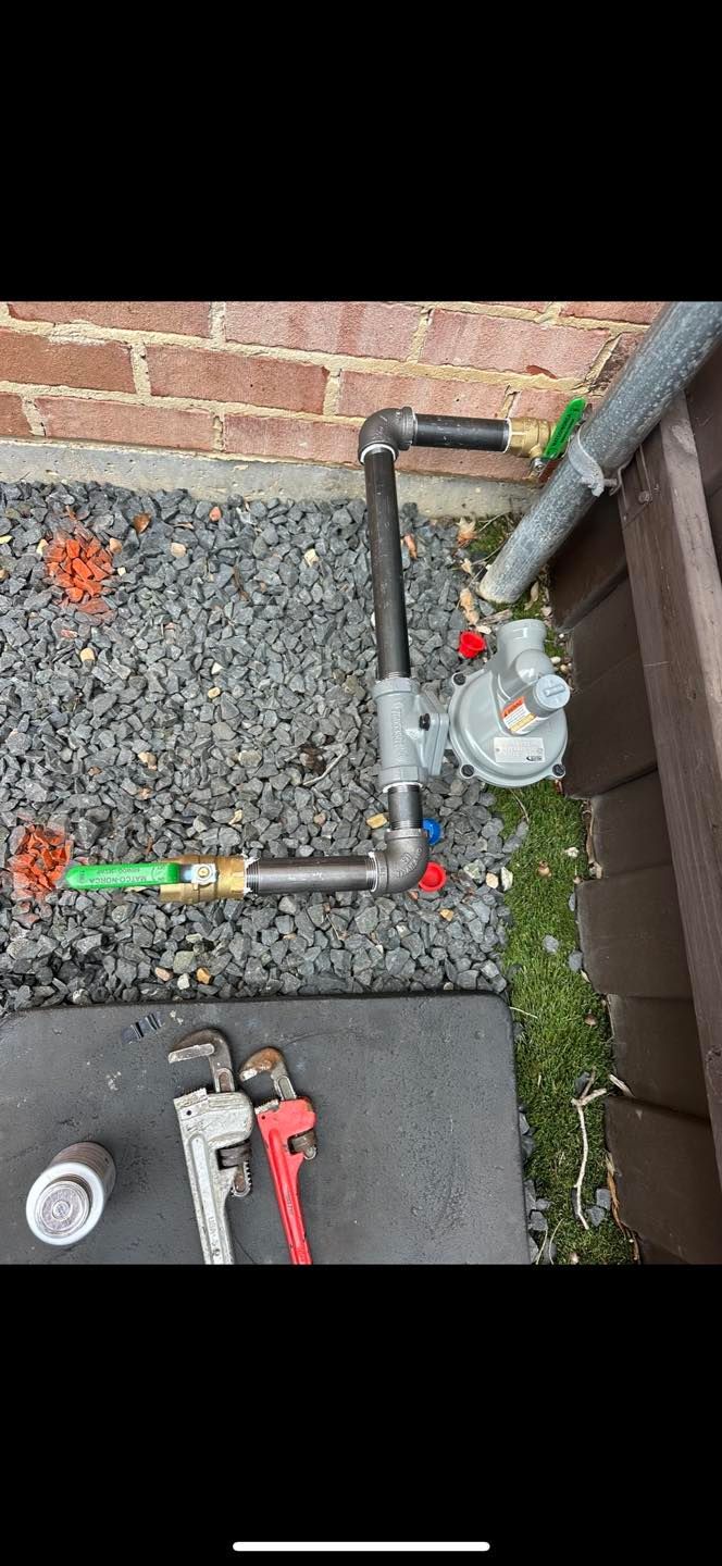 Outdoor utility pipes and a gas meter beside a brick wall, with tools on a paved surface.