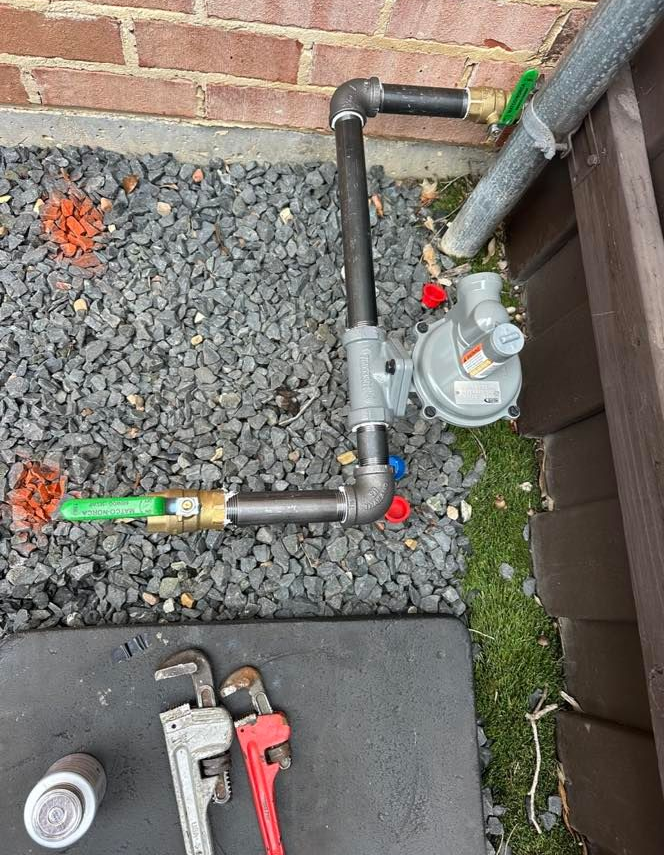 Water meter and pipes beside a brick wall, with a wrench on a sidewalk nearby.
