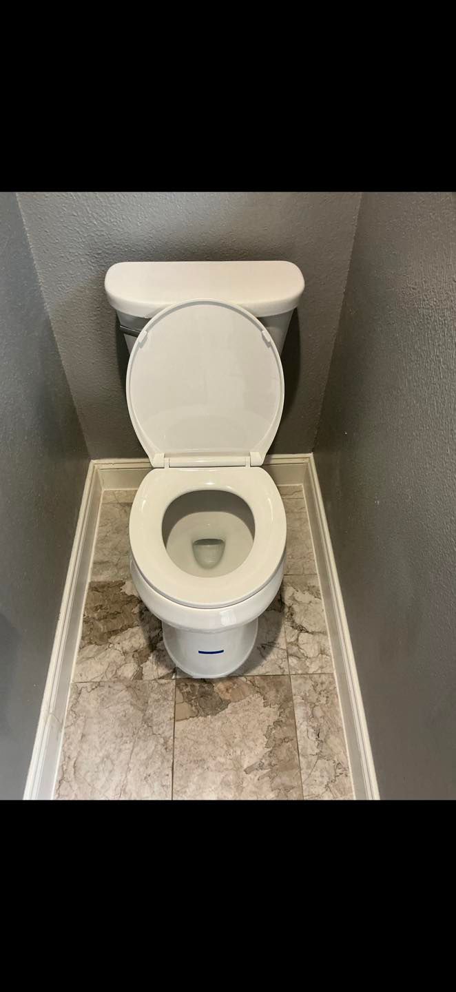 Open toilet in a small gray bathroom stall with a closed lid and textured floor mat.