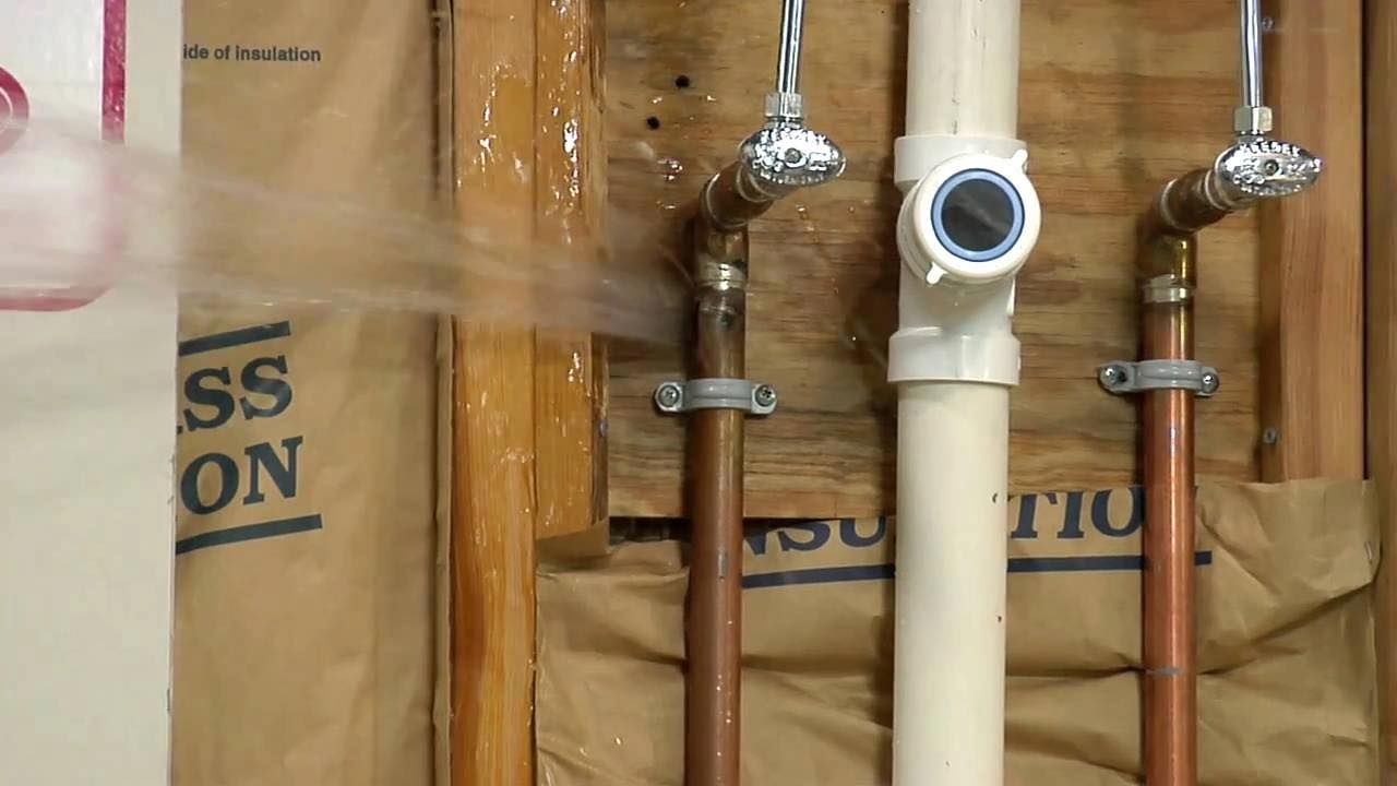 Plumbing pipes and valves in a wall cavity, including a white PVC pipe and copper lines.