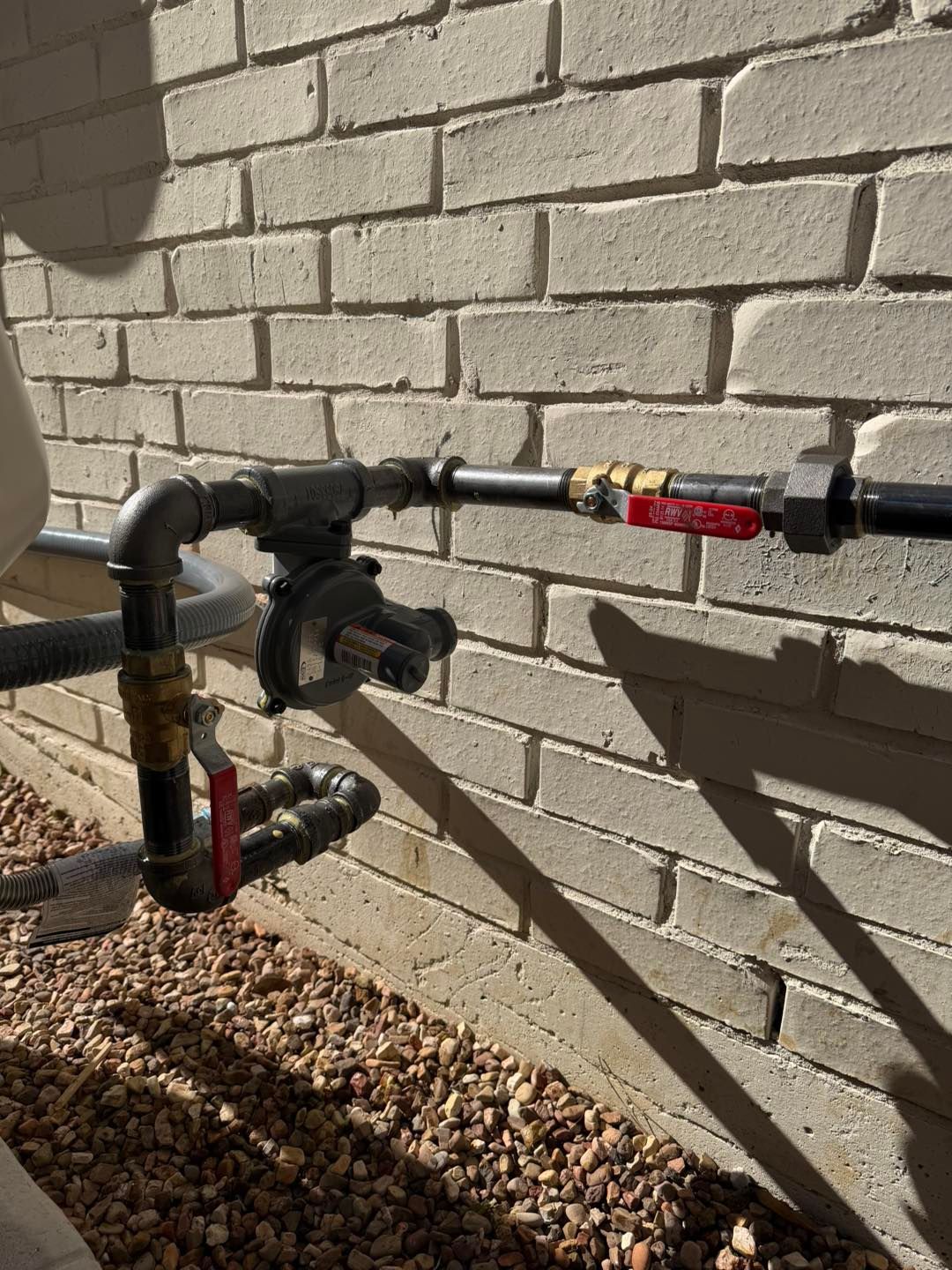 Outdoor gas meter and pipes mounted against a tan brick wall.