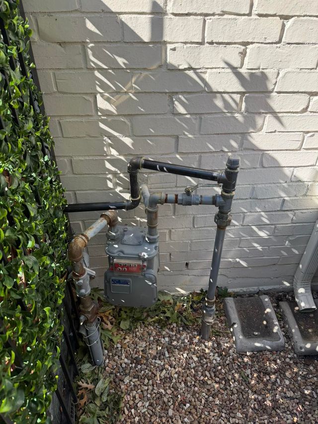 Gas meter and black piping mounted on a white brick exterior wall beside ivy and gravel.