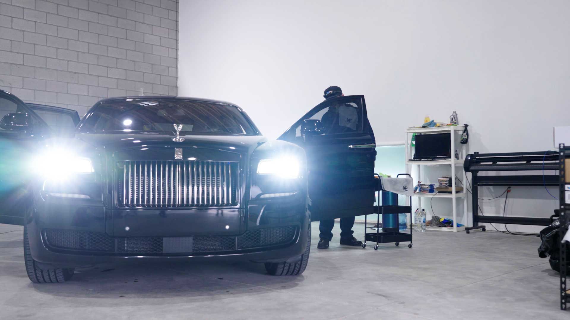 A person stands by an open door of a black Rolls-Royce with its headlights on inside a workshop.