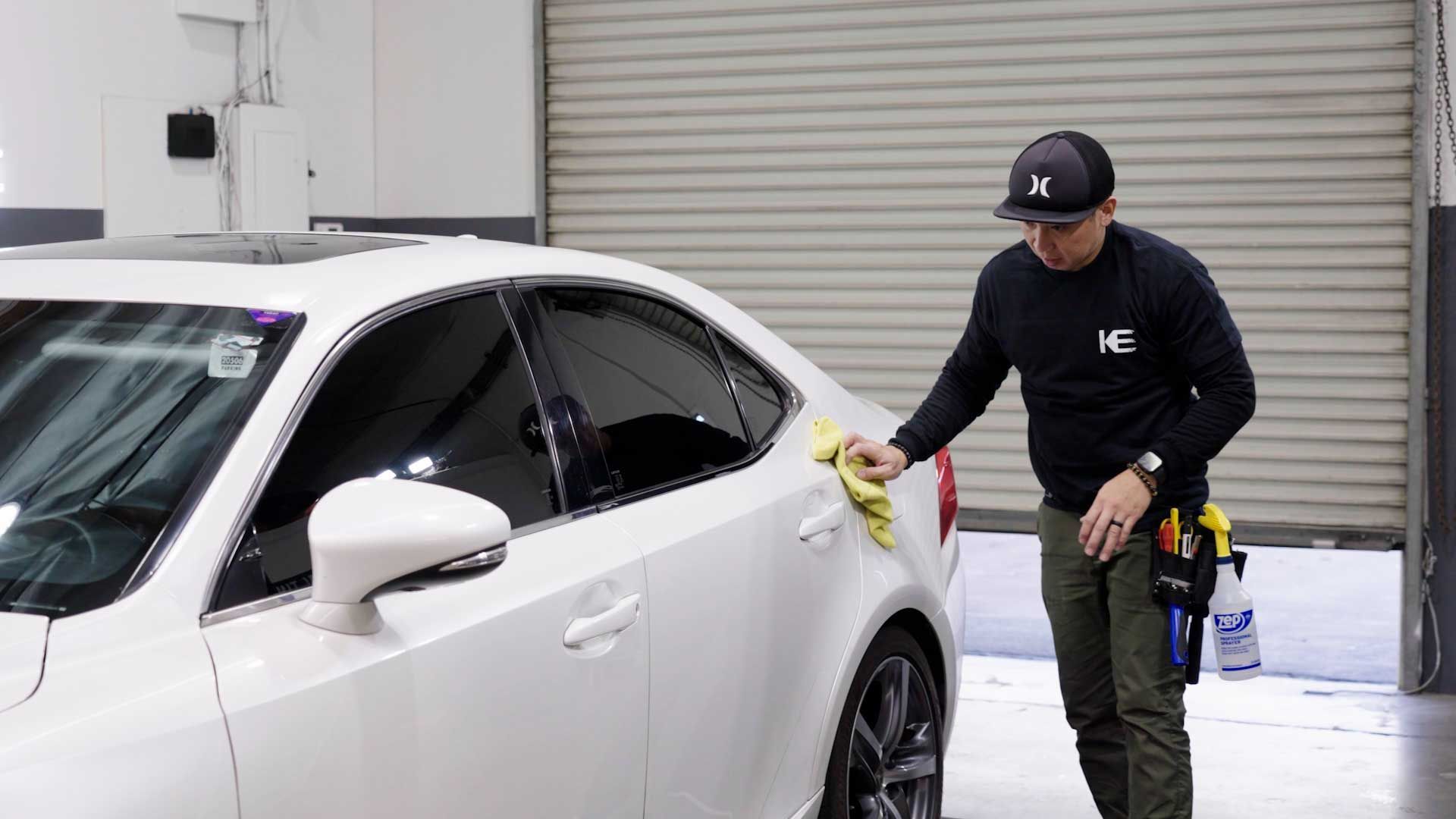 A person in a black shirt and cap wipes the white door of a sedan with a yellow cloth inside a garage.
