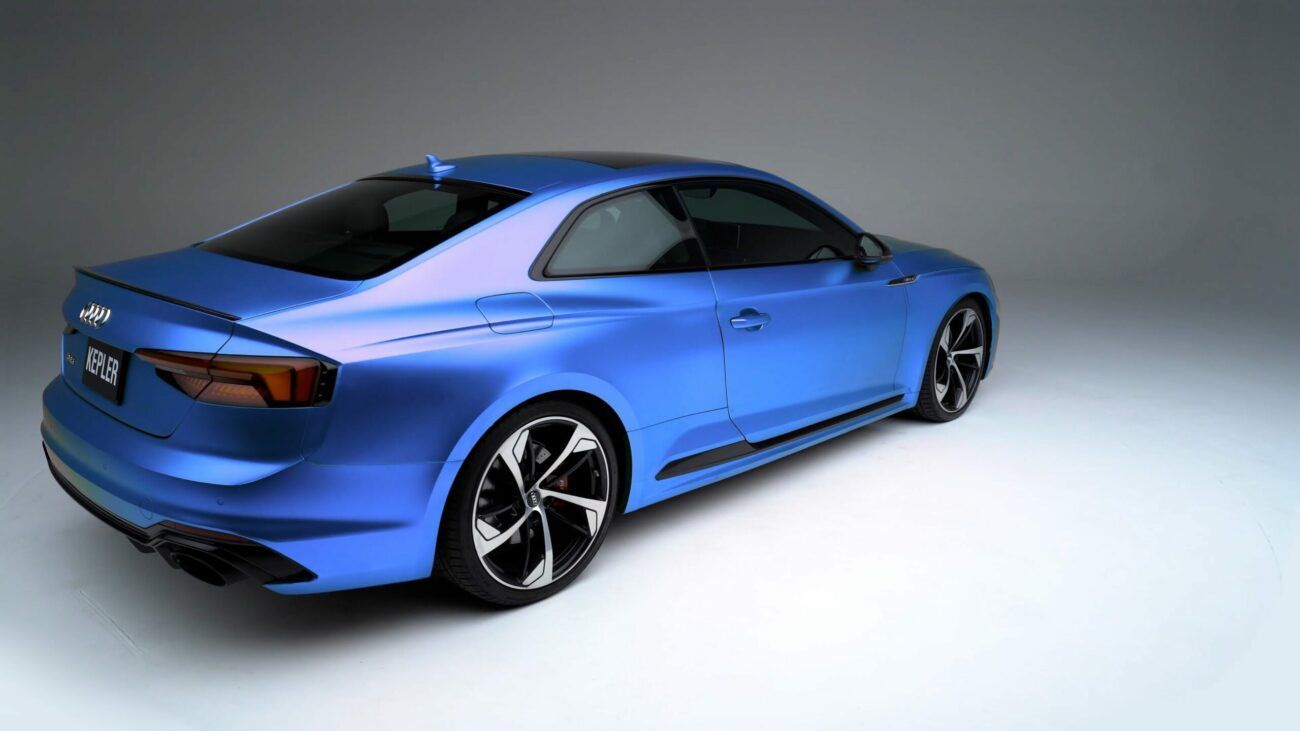 A blue Audi coupe seen from a rear three-quarter angle in a bright, minimalist studio setting.