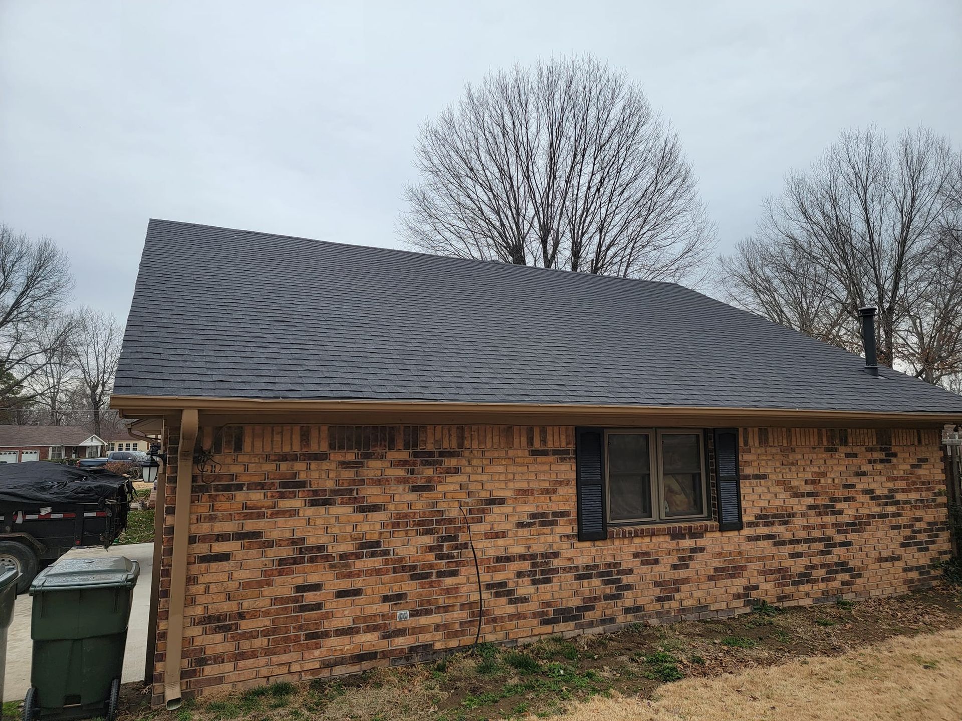 A brick house with a black roof and a green trash can in front of it.