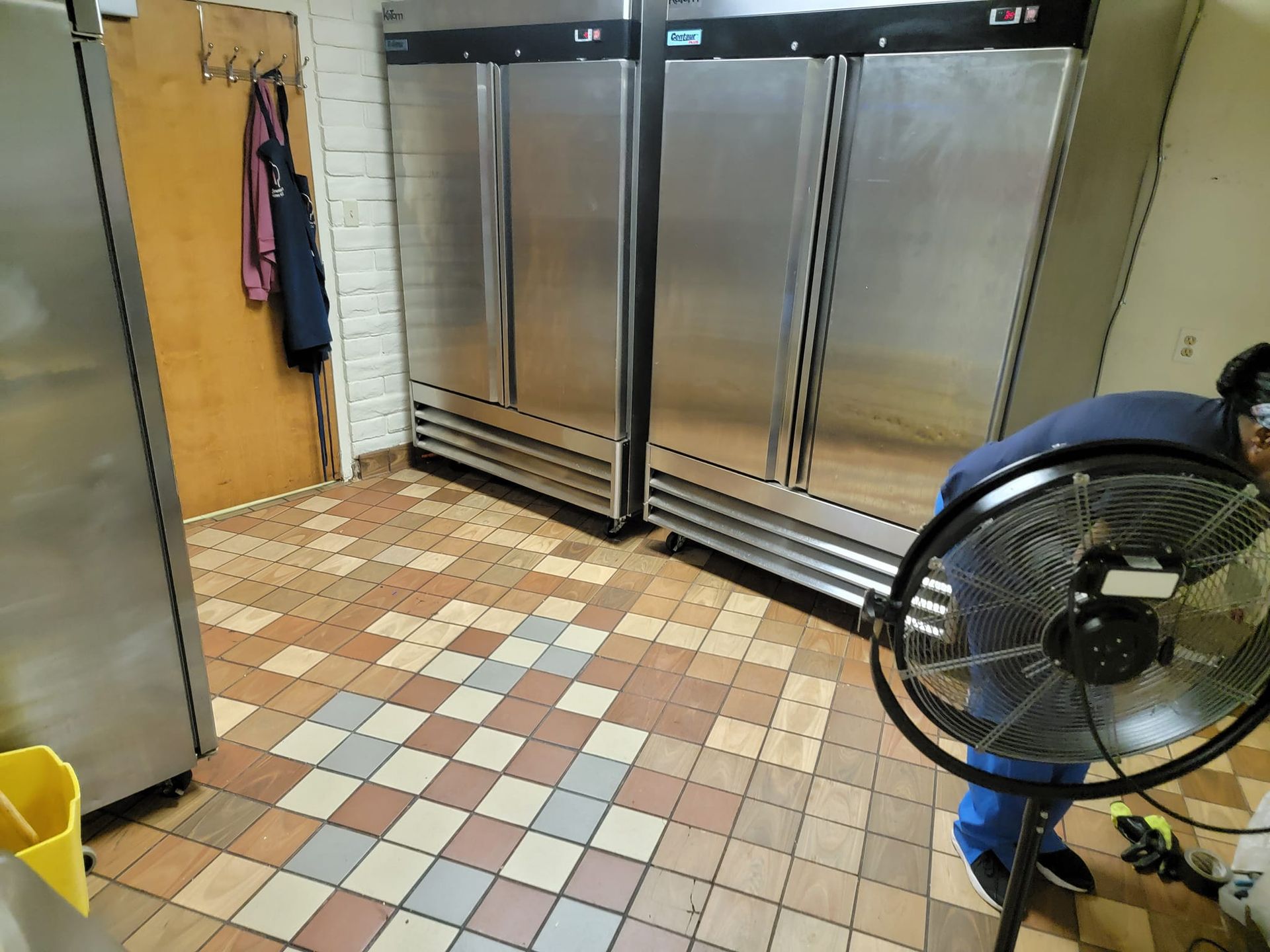 A man is standing next to a fan in a kitchen with stainless steel refrigerators.