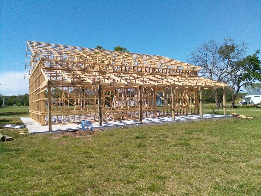 Wooden frame of a building under construction on a concrete foundation, in a grassy field under a blue sky.