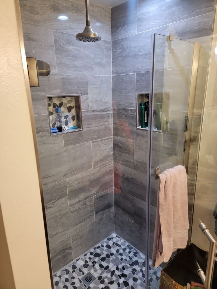 Grey tiled shower with glass door, shower head, recessed shelves, and pebble floor.
