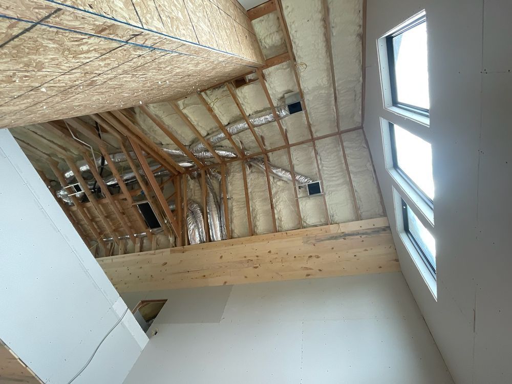 Interior view of a room under construction, showing open ceiling beams, insulation, and windows with sunlight.