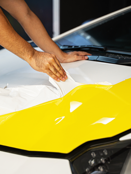 Hands smoothing white wrap onto a bright yellow car hood with the windshield in the background