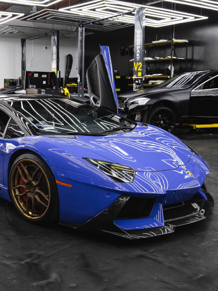 Blue Lamborghini with scissor door open in a garage, with a black sports car behind it.