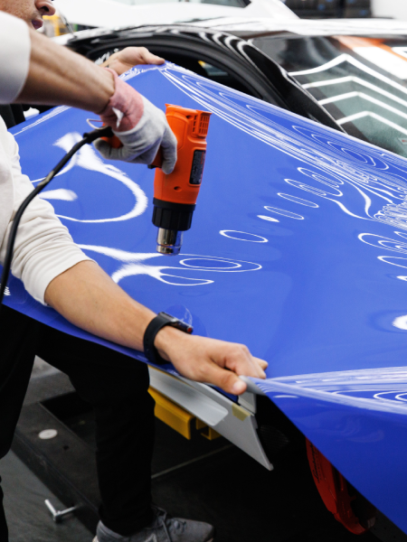 Technician using an orange heat gun on a blue car hood in a workshop