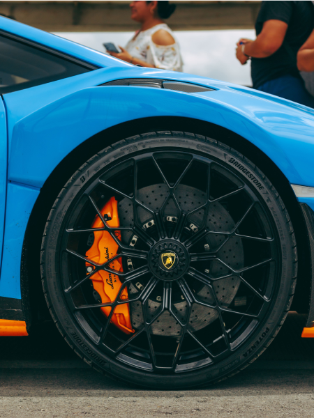 Blue sports car wheel with black rim and orange brake caliper, with people blurred in the background