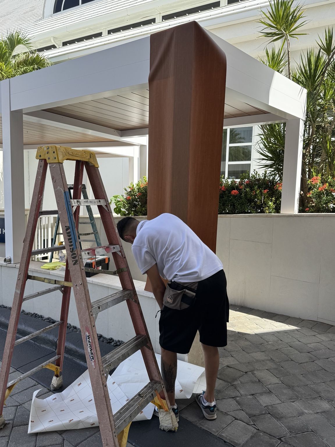 Person painting a wooden column beside a ladder under a covered outdoor patio