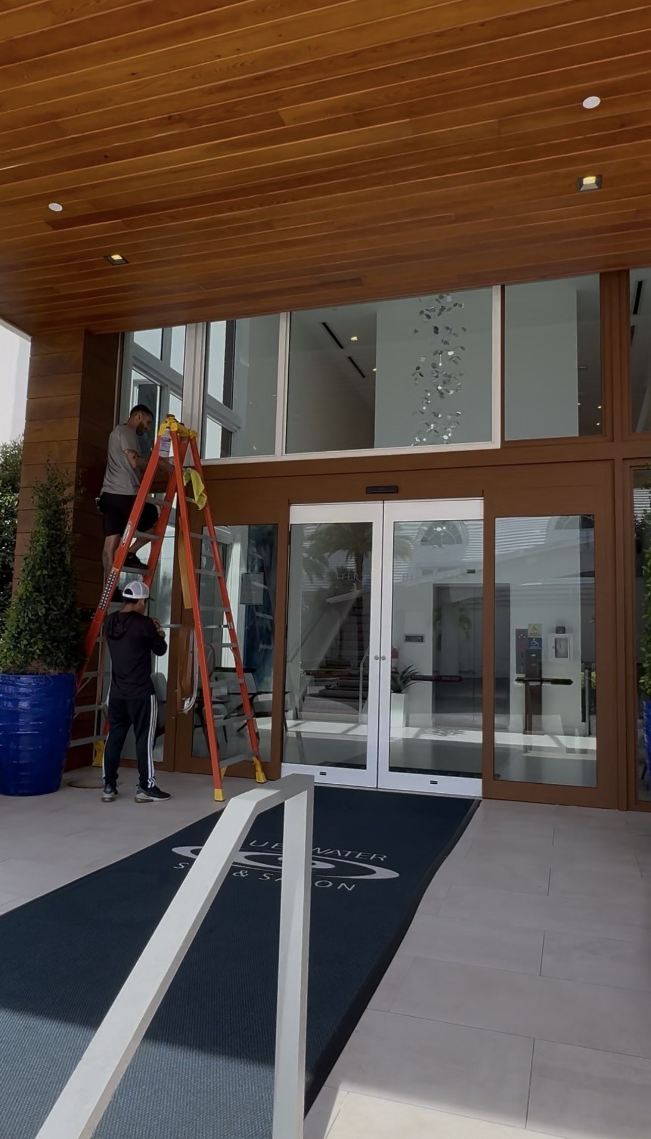 Person on a ladder outside a glass building entrance, under a wooden overhang.