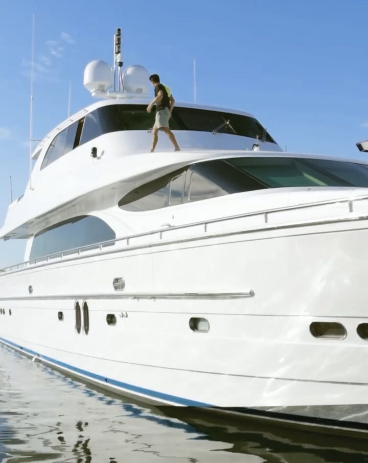 White yacht docked at a marina, with masts and boats reflected in the calm blue water.