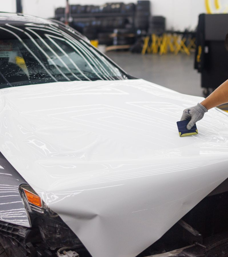 Man applying white vinyl wrap to the hood of a car in an auto workshop