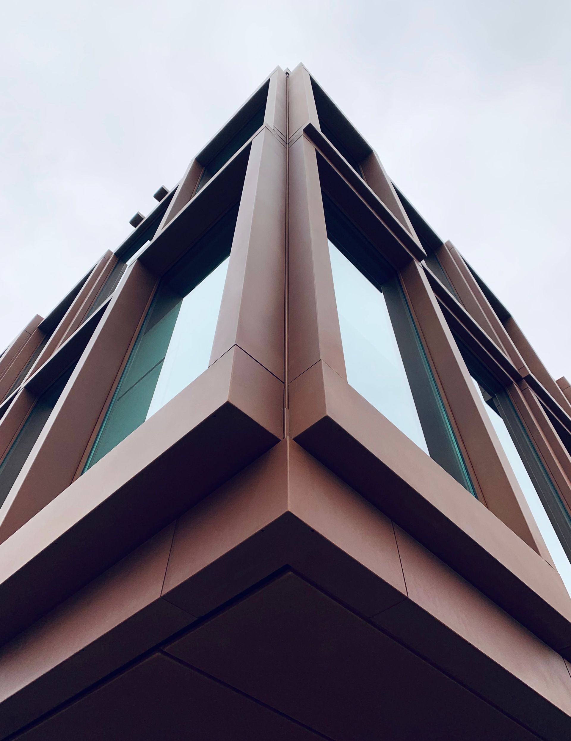 Low-angle view of a modern building corner with brown and teal panels against a cloudy sky