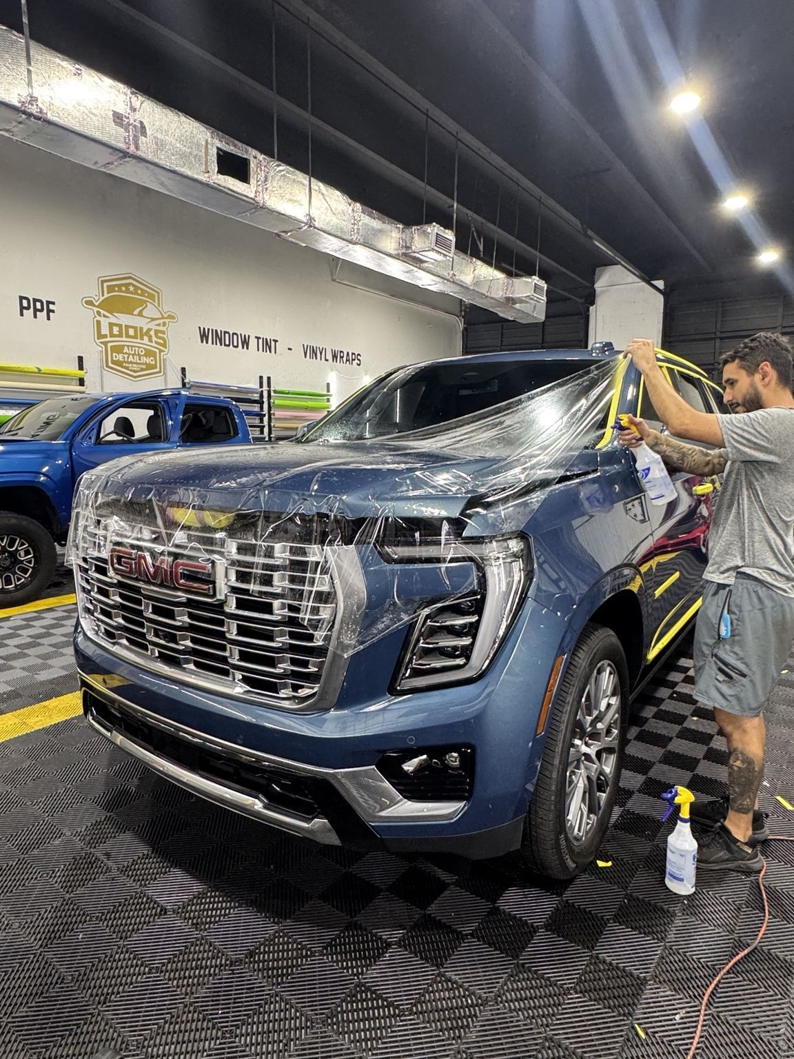 Blue pickup truck being washed indoors by a worker with a pressure washer and suds on the hood