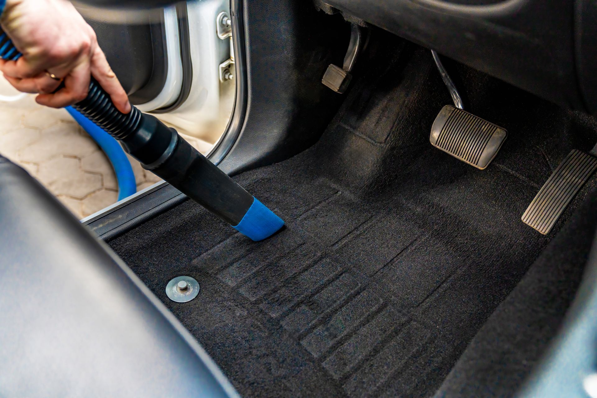Hand vacuum cleaning dirt from a car’s black floor mat near the pedals