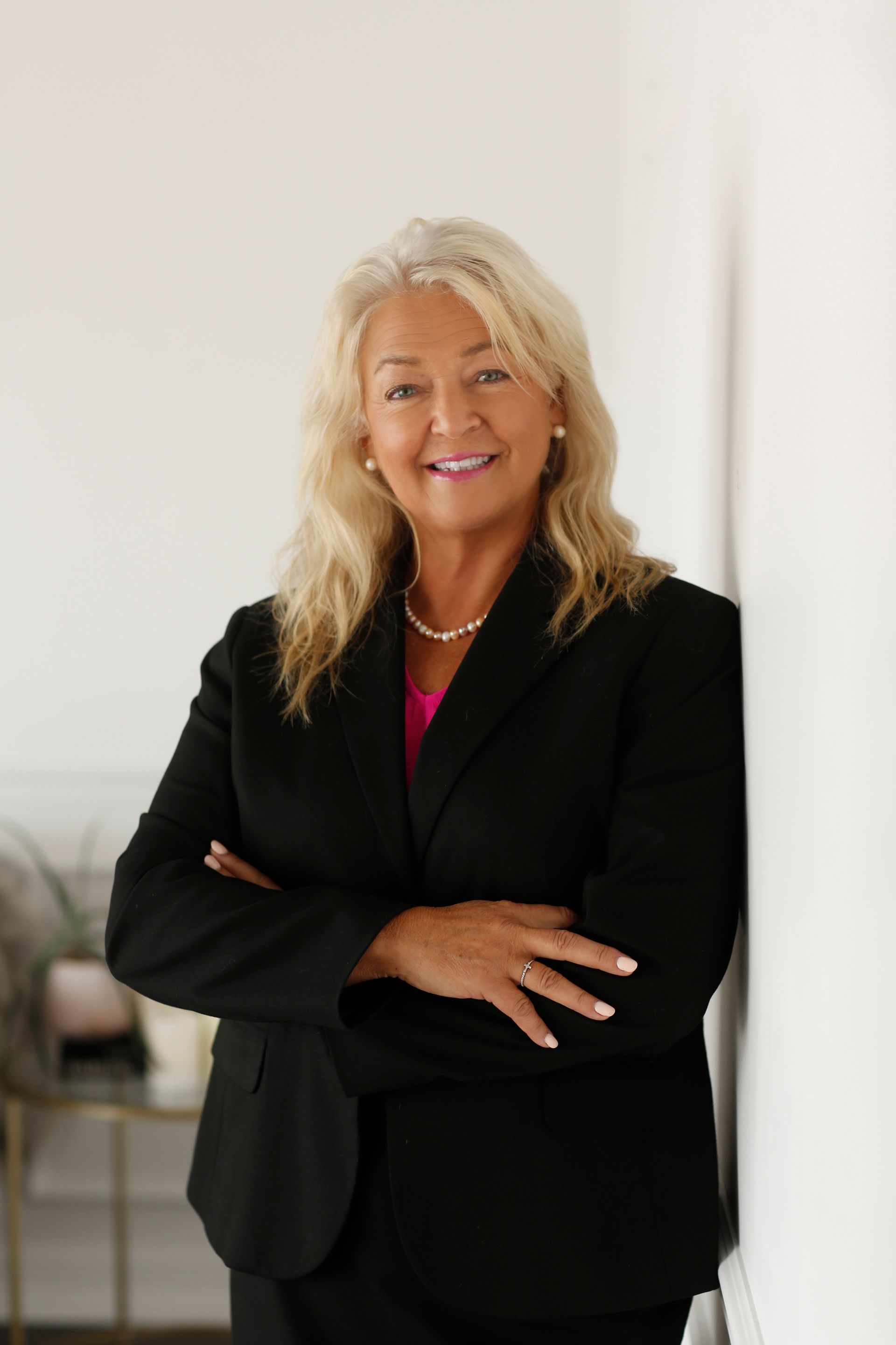 A woman in a black suit is leaning against a wall with her arms crossed.