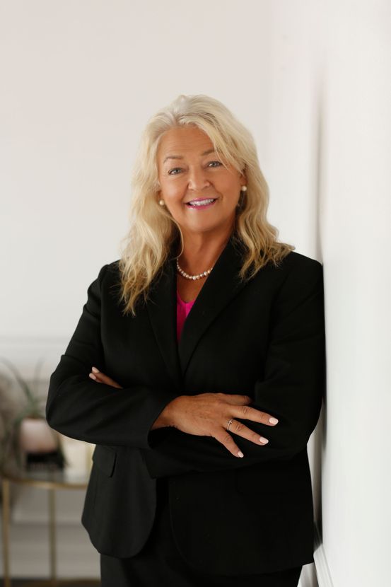 A woman in a black suit is leaning against a wall with her arms crossed.