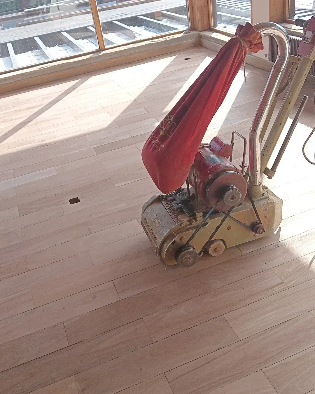 Un carrito de equipaje antiguo con una bolsa roja sobre un suelo de madera iluminado por el sol cerca de las ventanas.