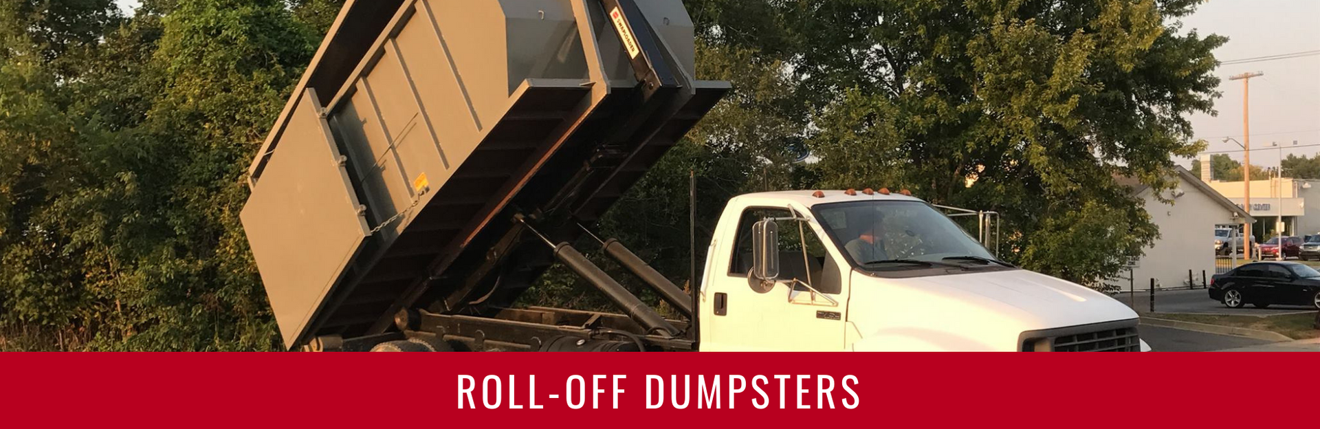 A white truck with a large, dark-colored roll-off dumpster tilted up at an angle against a backdrop of trees.