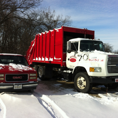 A white garbage truck with a large red container parked next to a red GMC pickup truck on a snowy, sunny day.