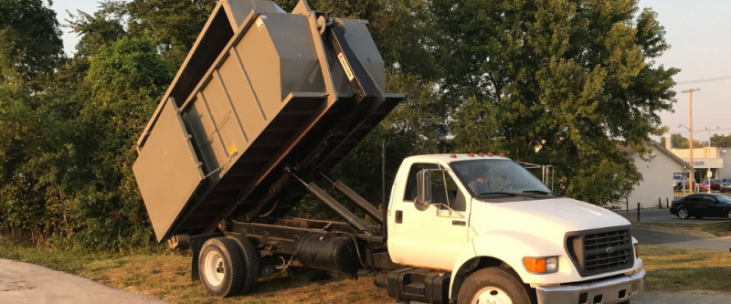 A white dump truck is parked on a dirt shoulder with its metal dumpster container tilted upward in an unloading position.