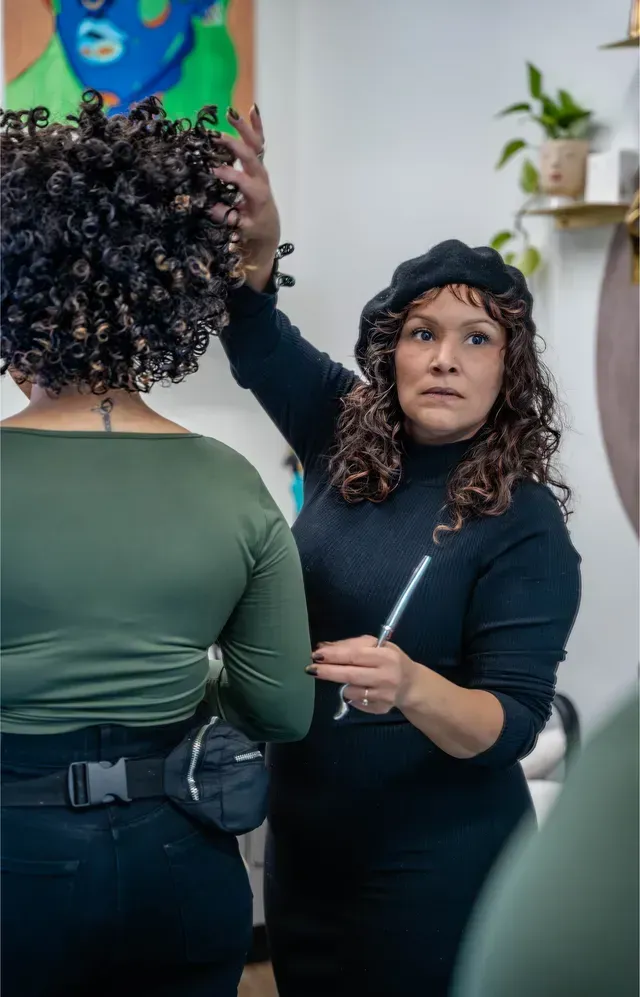 Stylist adjusting a curly-haired client's hair in a salon mirror, holding a comb and brush.