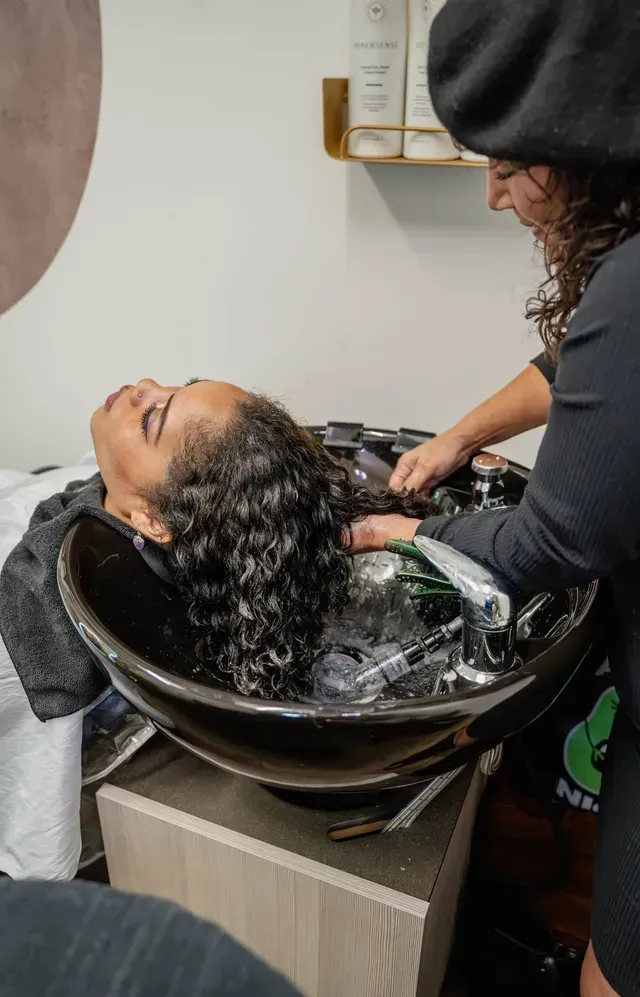 Salon sink hair wash with a stylist rinsing a client’s wet curly hair