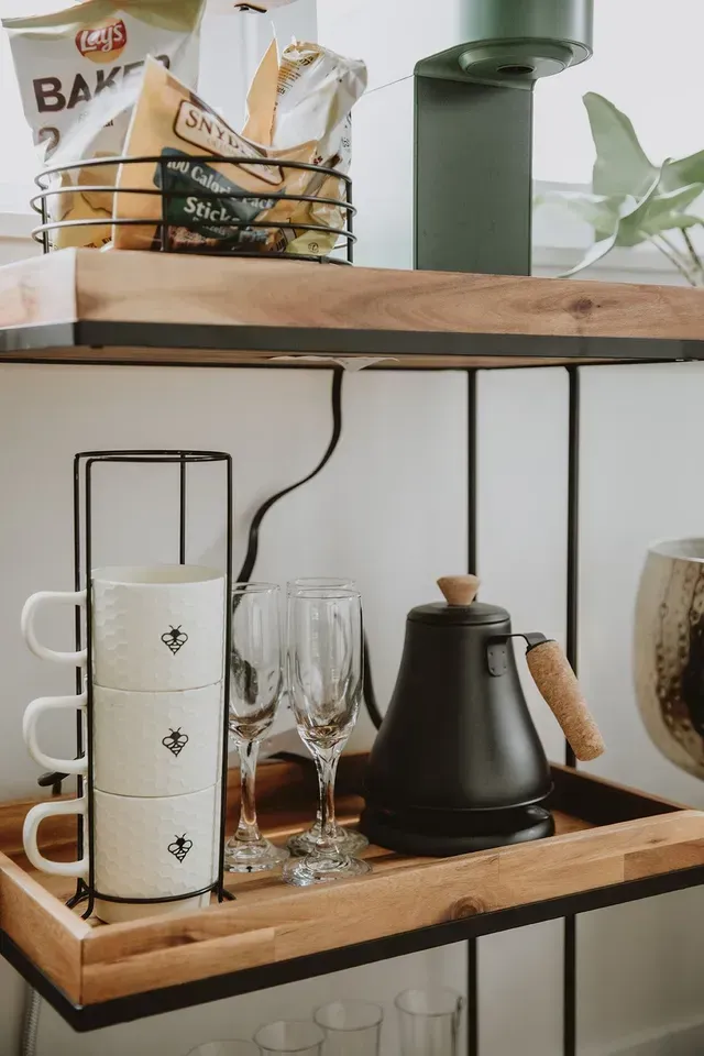 Wooden kitchen shelf with mugs, glasses, a black kettle, bread basket, and green decor
