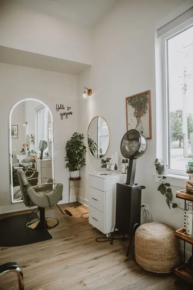 Bright minimalist salon with styling chair, mirrors, white dresser, plants, and large window.