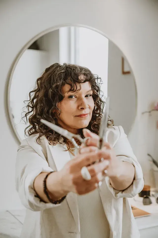 Person adjusting curly hair in a mirror, wearing a white top in a bright room