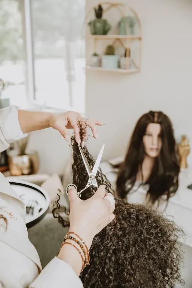 Hairdresser styling curly hair in a bright salon while a client sits in the background.