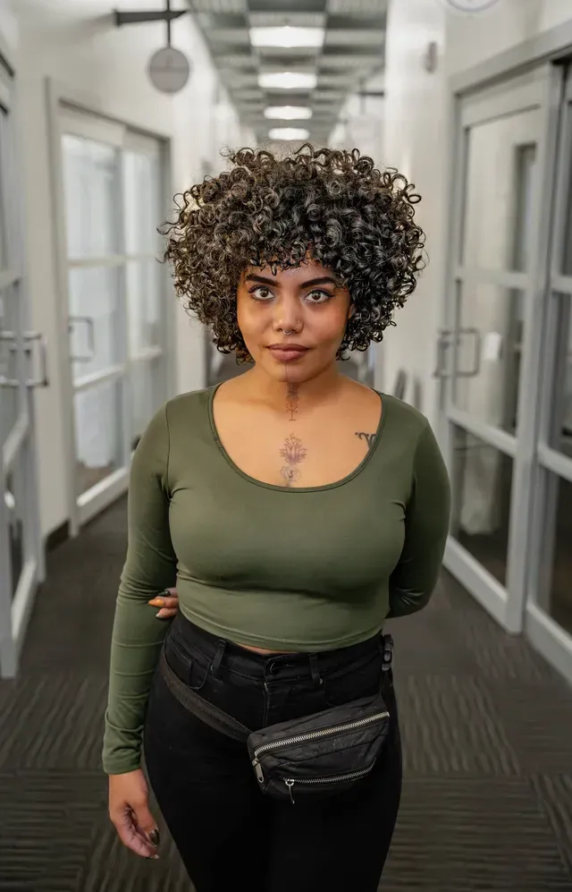 Person standing in a hallway, wearing an olive top and black crossbody bag, looking at the camera.