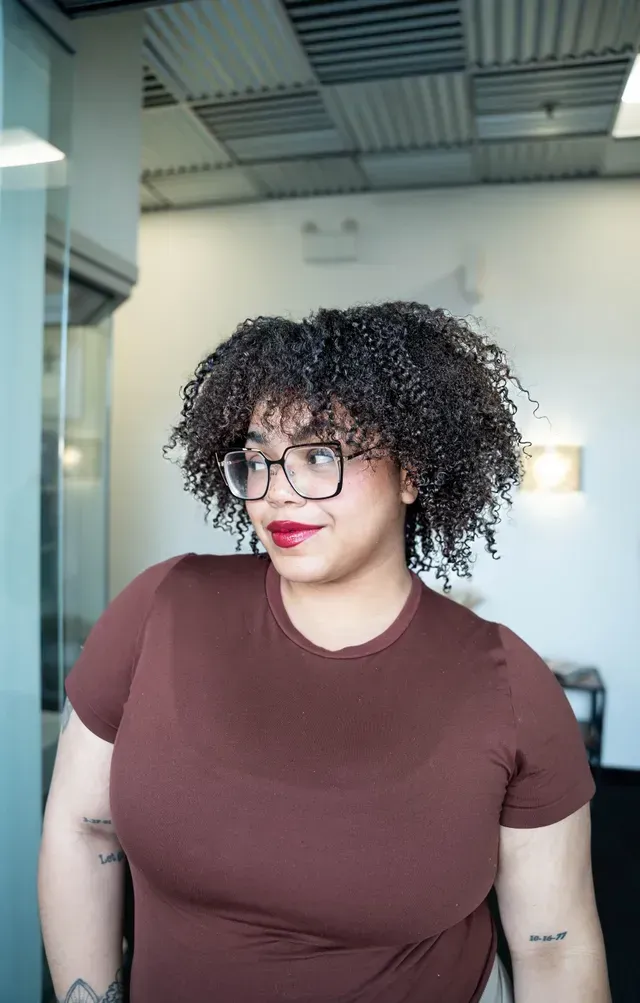 Person with curly hair and glasses wearing a brown shirt, standing indoors against a bright background