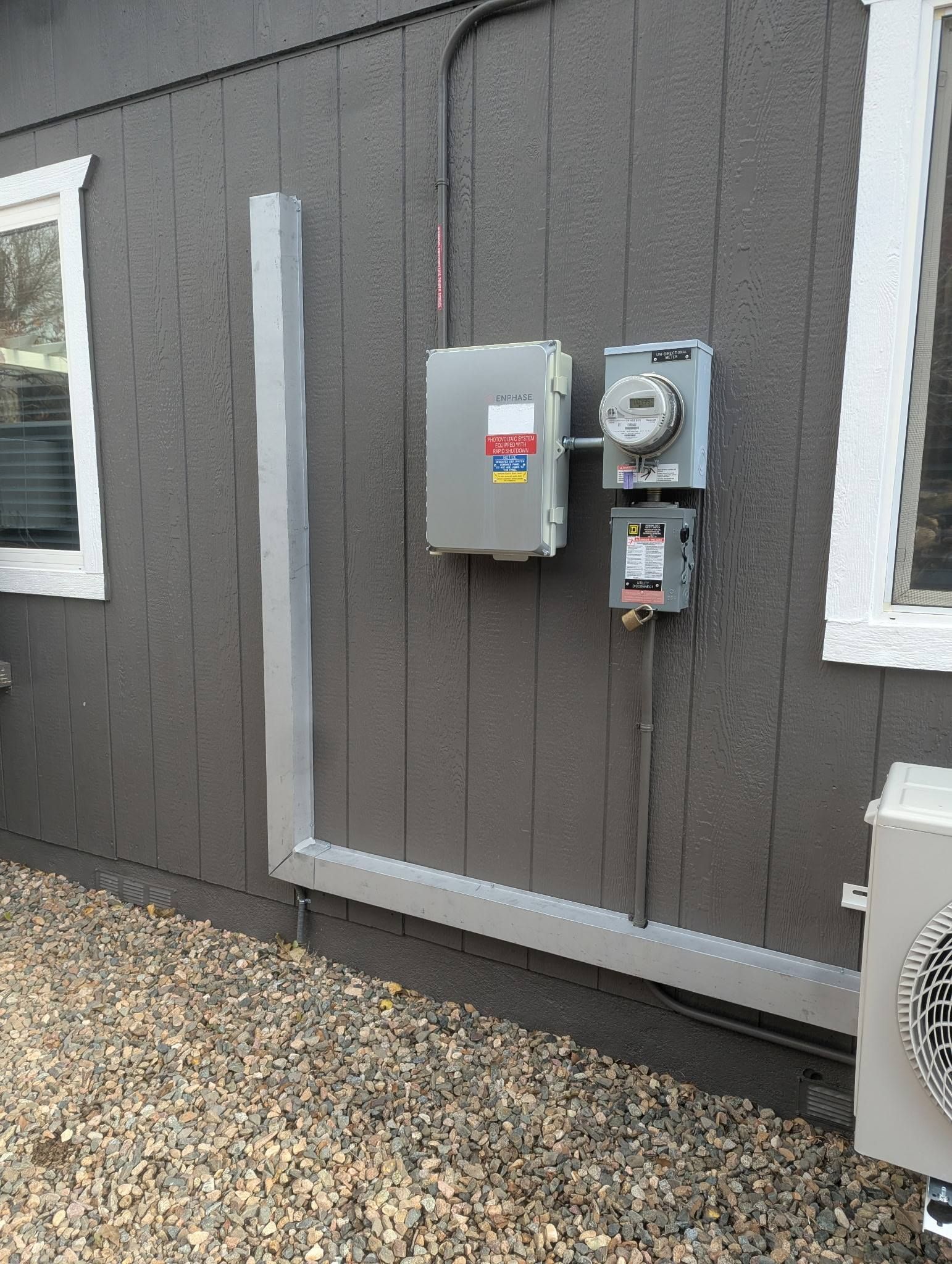 Exterior wall with utility meter and electrical boxes beside a window, over gravel.