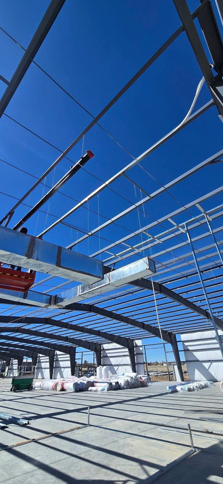 Construction site with steel roof frame under a bright blue sky, shadows across the ground