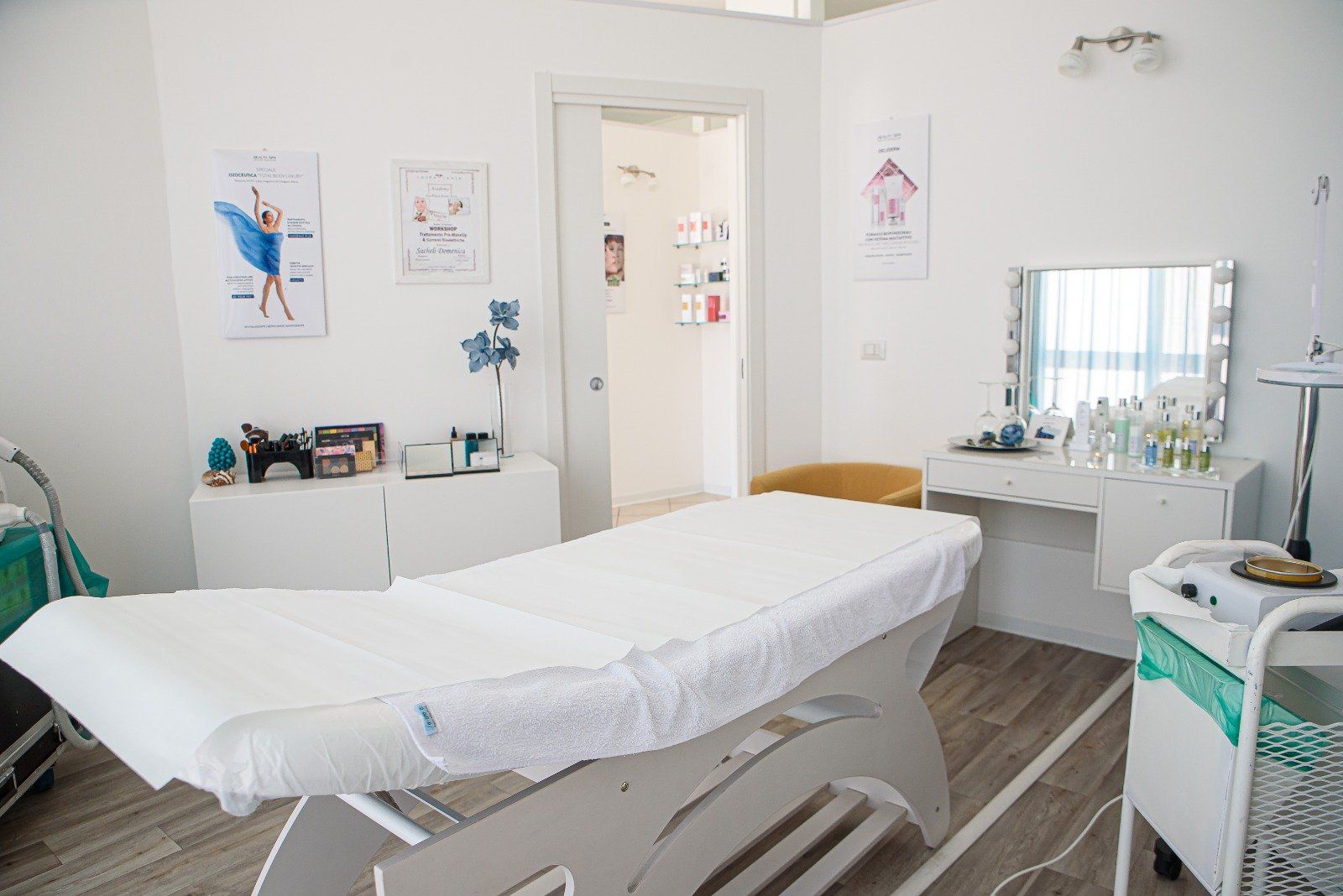 Sala spa con lettino per trattamenti, tavolo da trucco e poster. Luminosa e pulita.