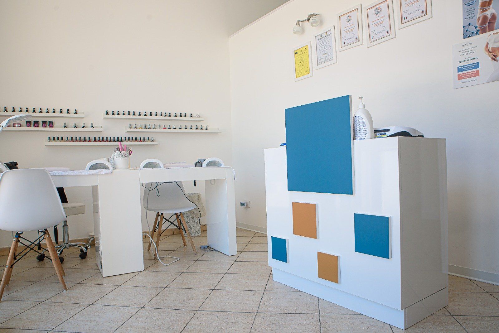 Interno di un salone per unghie con pareti bianche, tavolo per manicure, bancone della reception e scaffali per lo smalto.