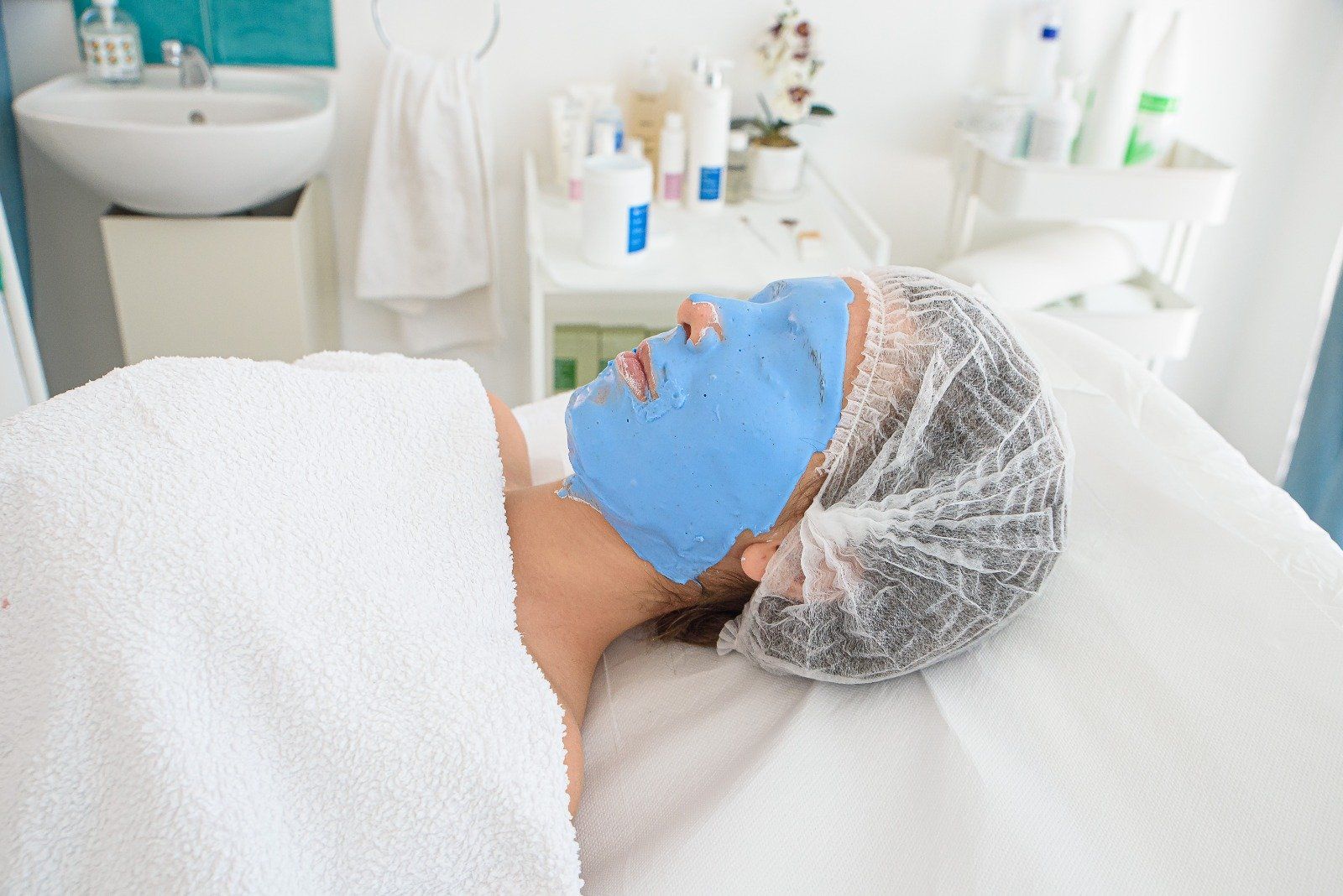Persona con maschera facciale blu, sotto un asciugamano bianco, sdraiata su un lettino da spa, con una retina per capelli.