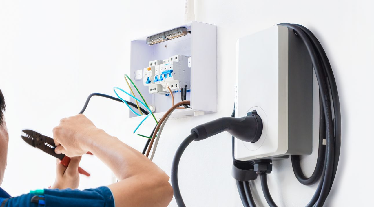 Two men are working on an electrical panel on a wall.