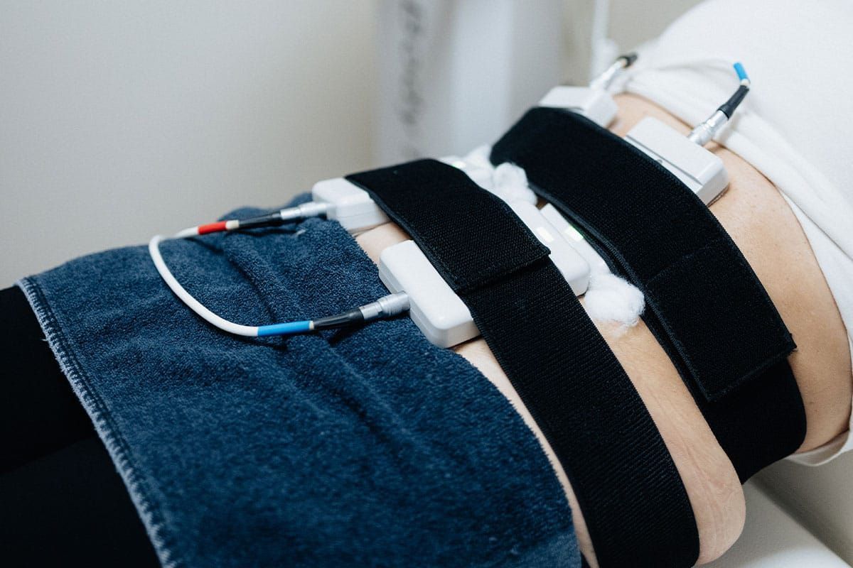 Person with electrodes attached to abdomen, wearing black straps, blue jeans visible.