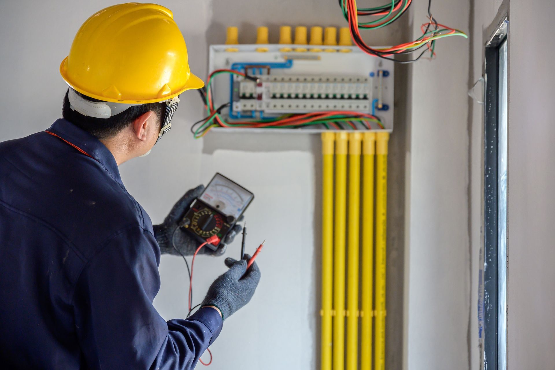 Electrician in hardhat using a multimeter to check a circuit breaker panel.