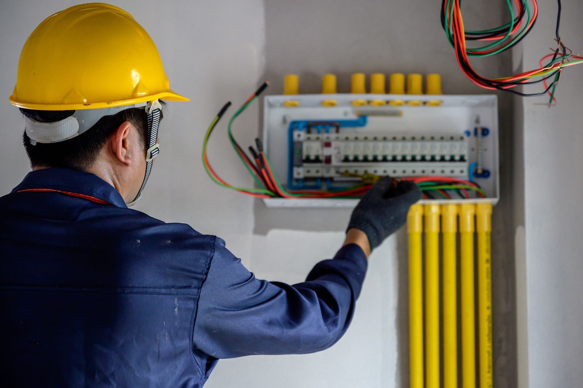 Electrician in blue uniform and yellow hardhat working on electrical panel, wiring.