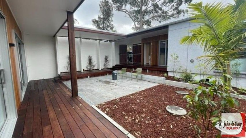 A modern courtyard with a wooden deck, stone patio, mulch garden bed, and light gray exterior walls.