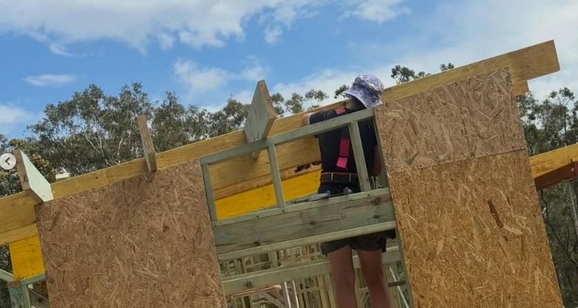 A construction worker in a sun hat stands on a wooden frame, installing exterior paneling on a building under a blue sky.